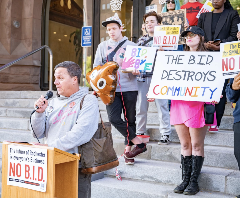 Gary Harding addresses the press standing behind a podium at the "Split the Bill" Rally on the steps of Rochester City Hall. There is a group of community members standing behind him with signs against the Business Improvement District in Downtown Rochester, NY because of the potential harm it will cause to the community. Photo by Jackie Photography.